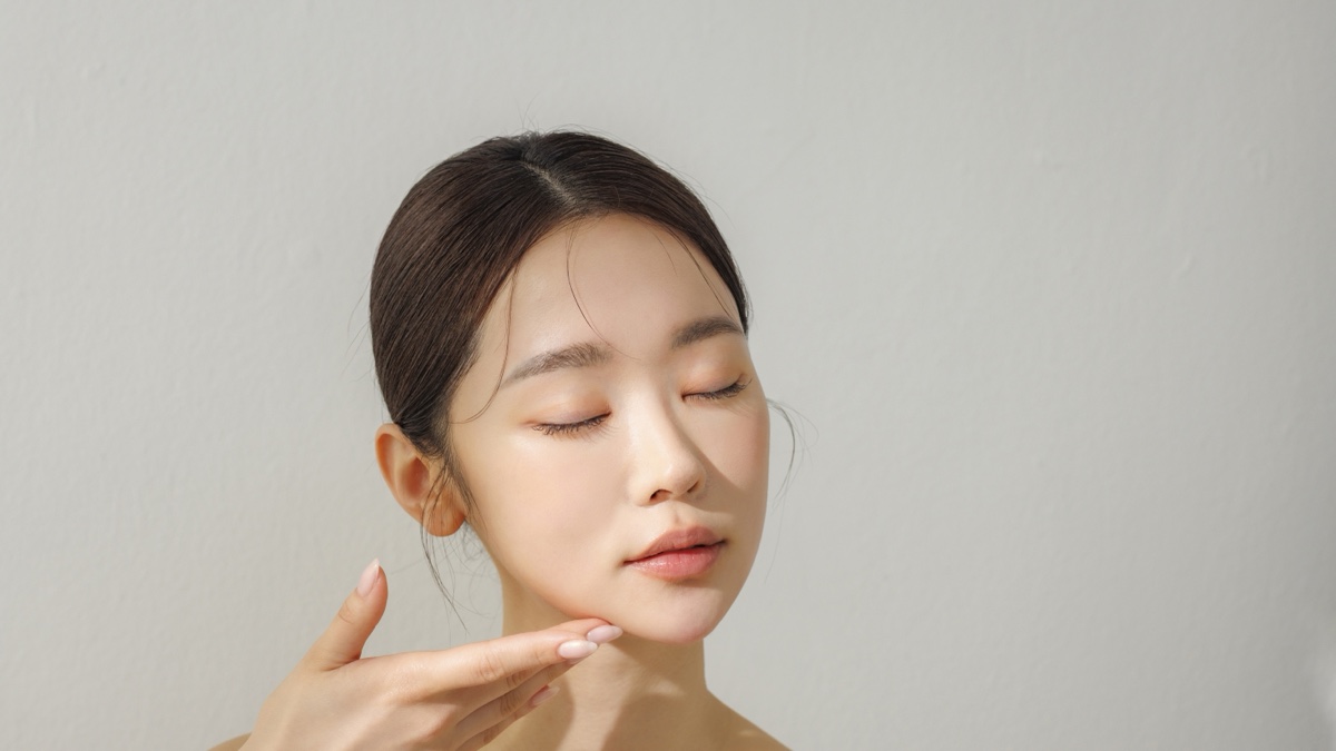 눈을 감은 여성이 손끝으로 턱선을 가볍게 받치고 있는 회색 배경의 뷰티 사진