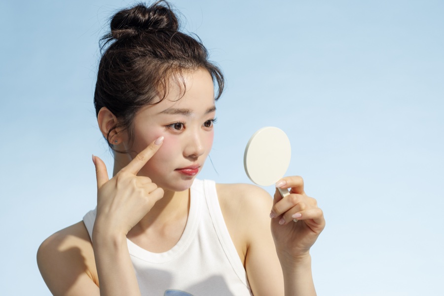 하늘색 배경에서 둥근 손거울을 들고 손가락으로 볼을 가리키며 피부 상태를 살피는 여성